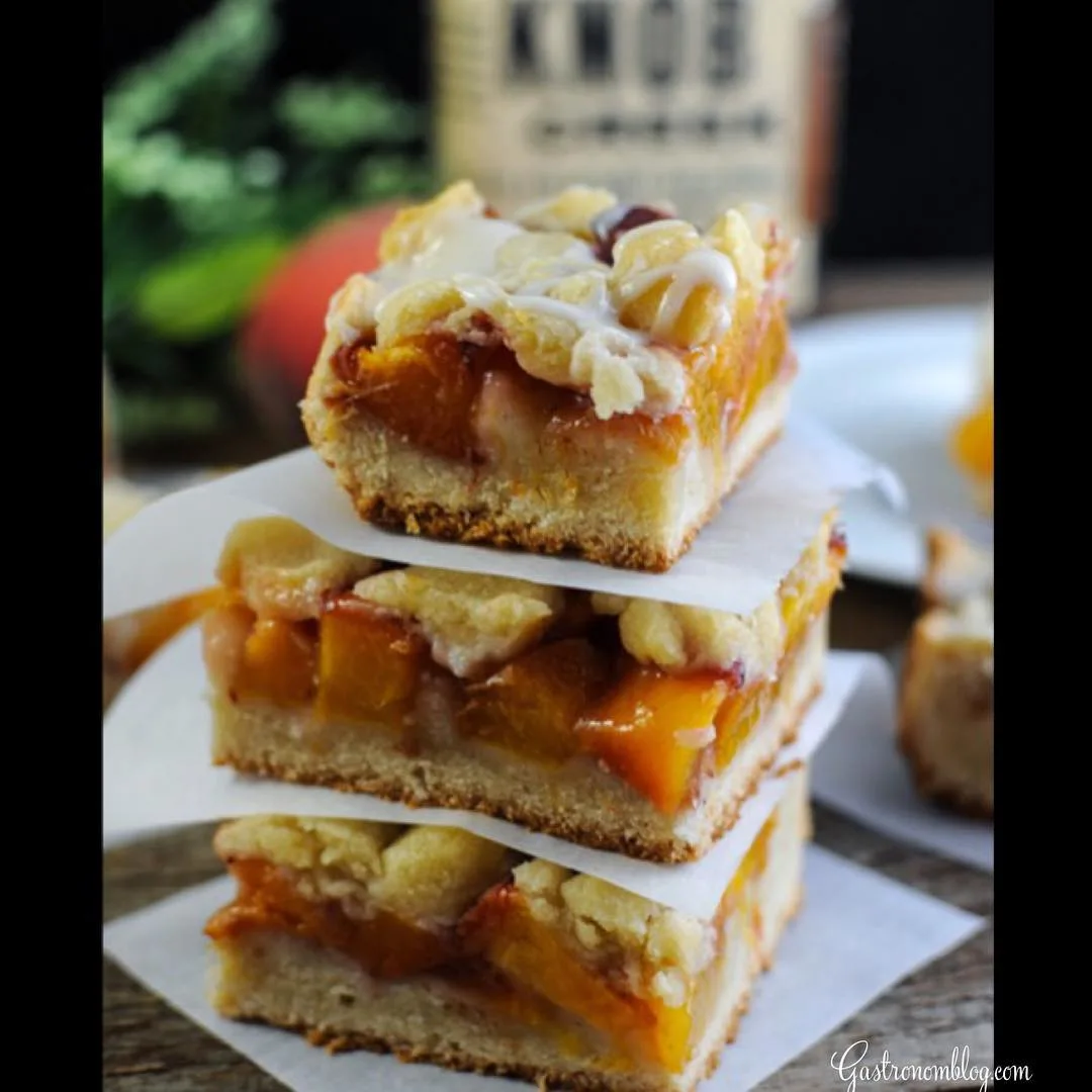 Peach bars stacked on each other separated by parchment squares. Whiskey bottle, peach and greneery in background