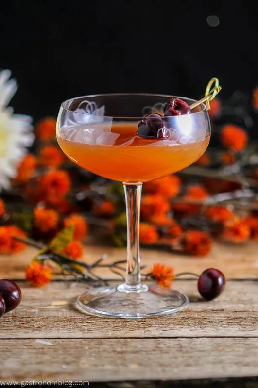 Apple Cider Manhattan, an orange cocktail in etched coupe with cherries on a bamboo pick. Orange flowers behind, on a wooden board with cherries