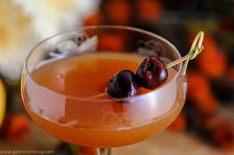 An Apple Cider Manhattan Cocktail in an etched coupe with brandied cherries on a cocktail pick with orange flowers in the background