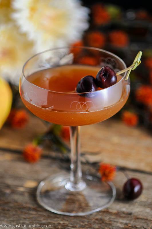 Apple Cider Manhattan - orange cocktail in a coupe with cherries on a pick. Flowers behind and on a wooden table. 
