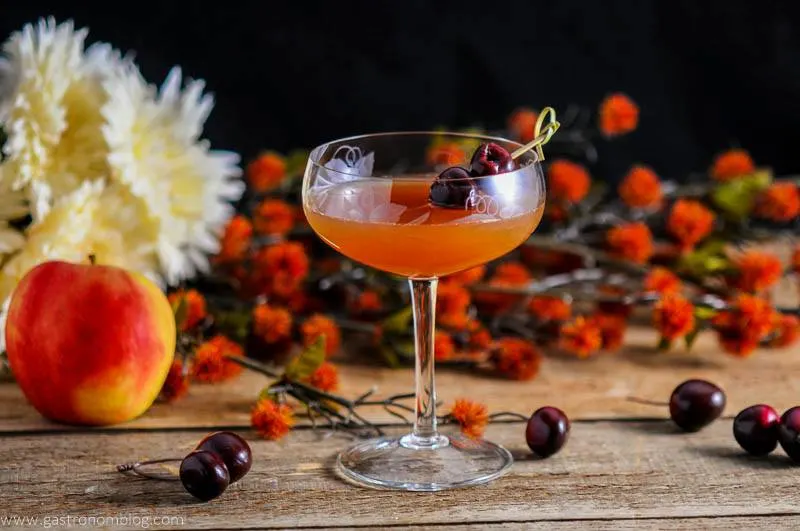 An Apple Cider Manhattan Cocktail in an etched coupe with brandied cherries on a cocktail pick with orange flowers in the background
