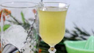 Yellow cocktail in tulip glass, greenery, glass mixing glass, silver jigger and green glass juicer behind