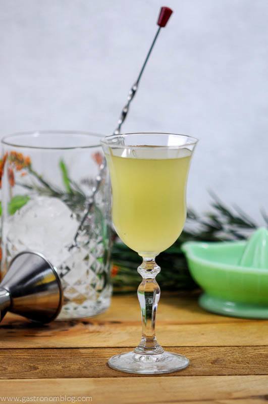 Yellow cocktail in tulip glass, greenery, glass mixing glass, silver jigger and green glass juicer behind