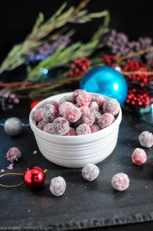 Sugared Cranberries in a white bowl, greenery and Christmas ornaments behind