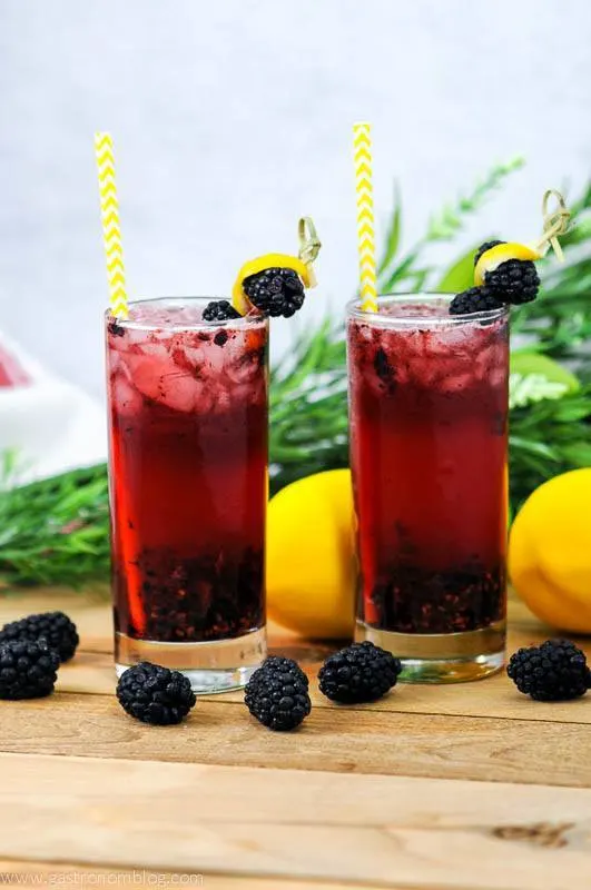Muddled Blackberry Gin cocktai in two highball glasses with yellow straws. Garnished with blackberries and lemon peel. Flowers in the background.