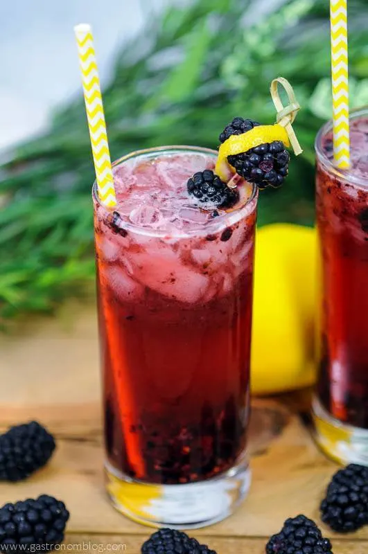 Muddled Blackberry Gin Cocktials in a highball glass with blackberries on table and cocktail pick. Yellow and white striped straws, greenery and lemons behind.