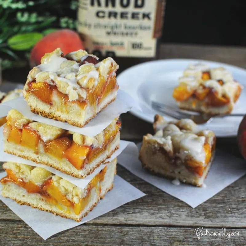 Stack of peach bars, one on plate and one on parchment paper. Whiskey bottle, peach and greneery behind