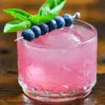 Pink cocktail in glass with blueberries on pick and basil