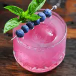 Pink cocktail in glass with blueberries and basil garnish