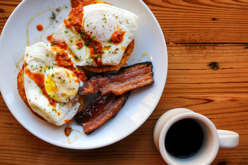 Top shot of eggs and bacon on white plate with hot sauce, coffee in white mug nearby