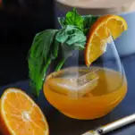 Orange cocktail in glass with slice of orange and basil, sliced orange and knife, gray container behind