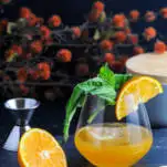 Orange cocktail in glass with orange slice and basil bunch, cut orange and knife with orange flowers behind