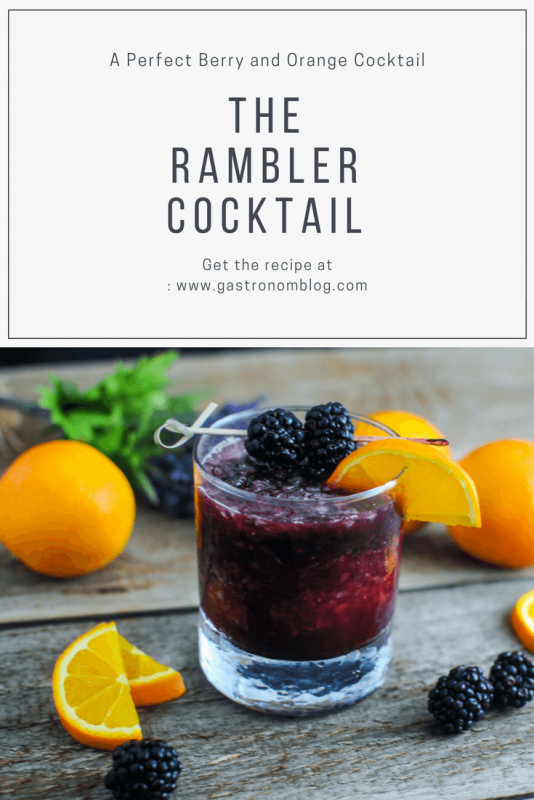 The Rambler Cocktail in rocks glass with berries on a cocktail pick with a slice of orange. Oranges and blackberries around glass. 