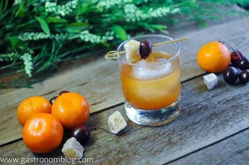 Ginger Clementine Old Fashioned Cocktail in a rocks glass with candied ginger, cherries and clementines on a wooden board