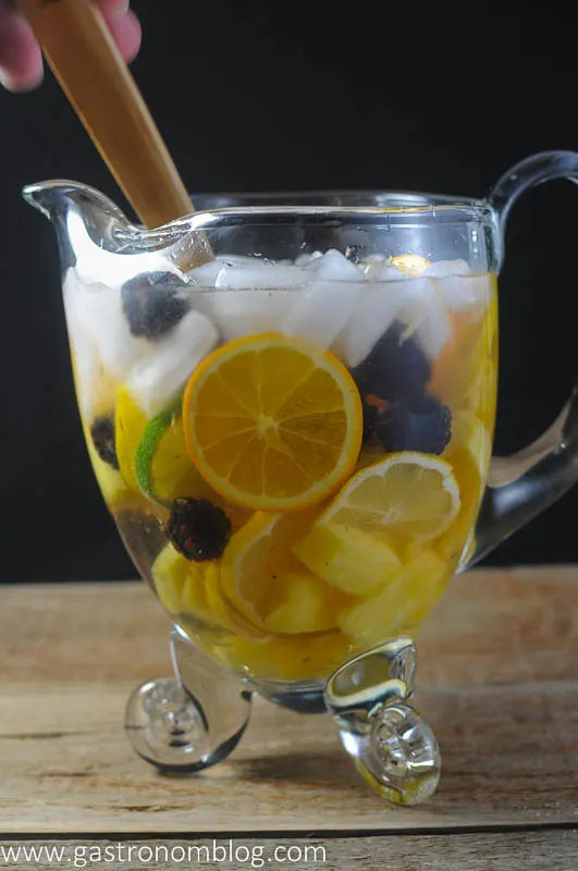 Wood spoon stirring in a clear pitcher filled with ice, white wine, blackberries, pineapple, lemons and oranges