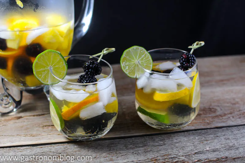 Two wine glasses filled with white sangria with limes, blackberries, oranges, lemons and pineapples. Pitcher in background
