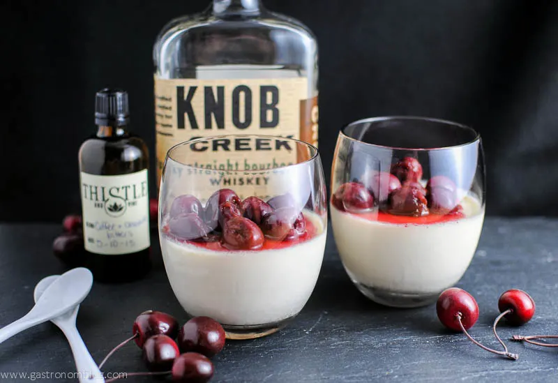 Old Fashioned Cocktail Panna Cottas in wine glasses, bitters and bourbon bottle in background with white spoons. Topped with cherries