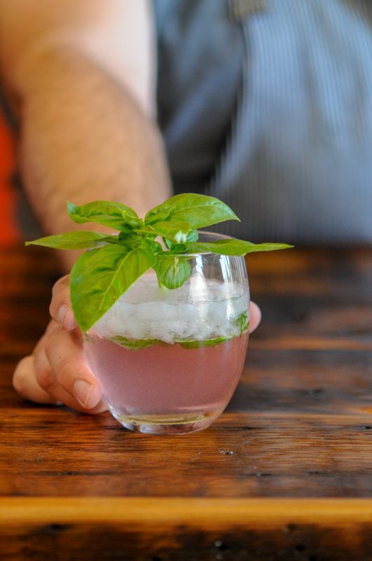 Rhubasil cocktail in glass with a bunch of basil, hand holding glass on wood table