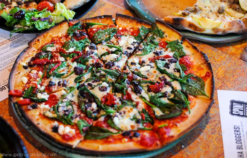 Pizza with fresh spinach, veggies, tomatoes and cheese on top. Salad and menu in background. 