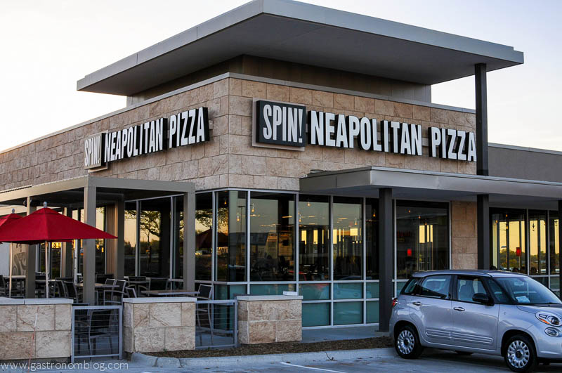 Picture of outside of Spin! Neapolitan Pizza restaurant in Omaha Nebraska - tan stone and patio with red umbrellas. Silver car in parking lot. 