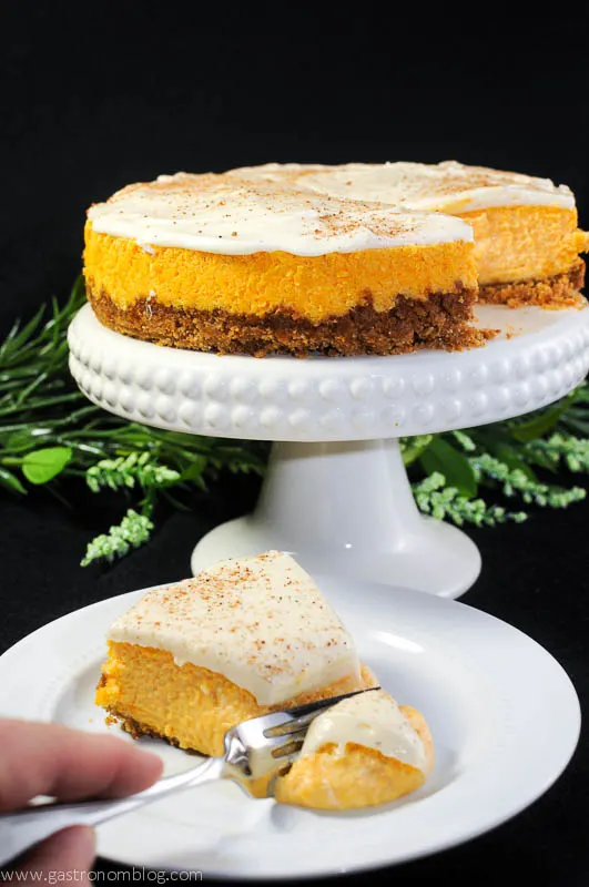 Carrot Cake cheesecake on a white cake stand and a slice being cut on a white plate. Flowers in background