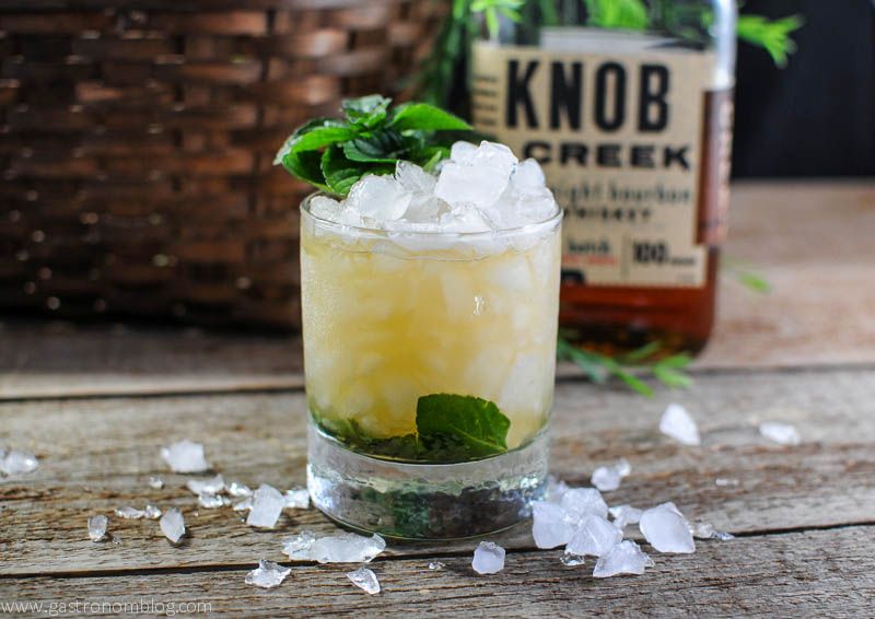 Mint Julep for the Kentucky Derby in a rocks glass with crushed ice and mint leaves. Basket and Knob Creek bottle in background. 