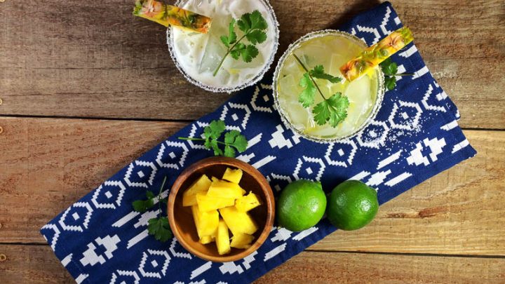 Top shot of yellow margaritas in rocks glasses with pineapple slices and cilantro. Glasses and wooden bowl of pineapple chunks with limes on blue and white napkin on wooden table