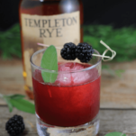 Red cocktail in glass with whiskey bottle behind, blackberries on pick, sage leaves