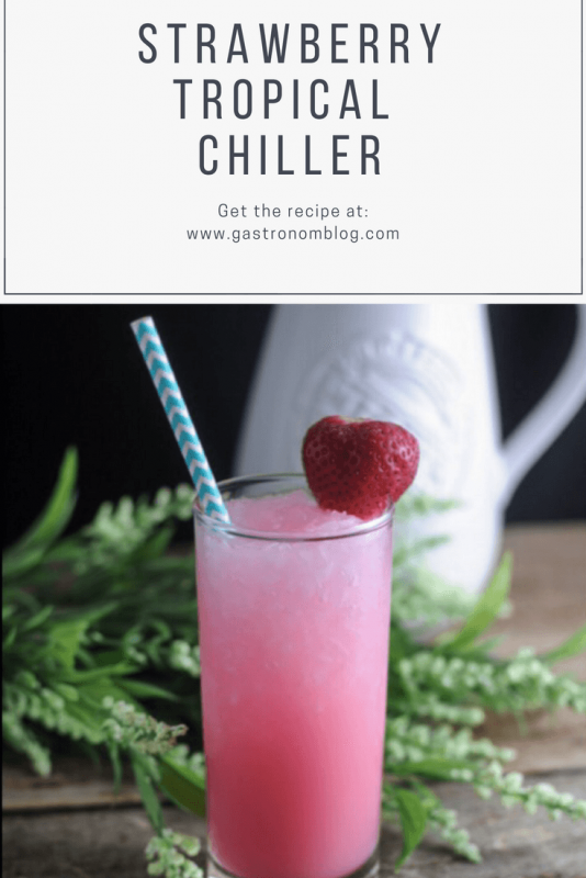 Strawberry Tropical Chiller Cocktail - pink cocktail in a tall glass with blue/white striped straw and a strawberry. Greenery and white pitcher in background
