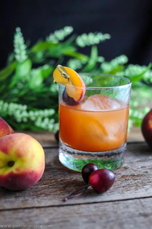 orange cocktail in rocks glass with round ice ball. Peaches, cherries on cocktail pick and next to glass. Flowers behind glass