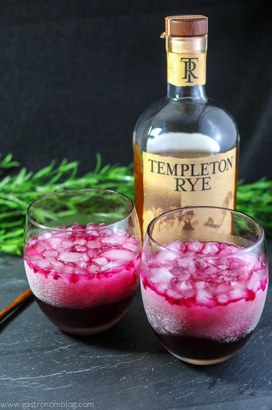 Red Beet Shrub Cocktail in two wineglasses, wooden spoon and whiskey bottle in background