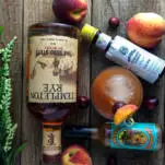 Top shot of peach cocktail and ingredients with whiskey bottle, bitters, peachs and hot sauce