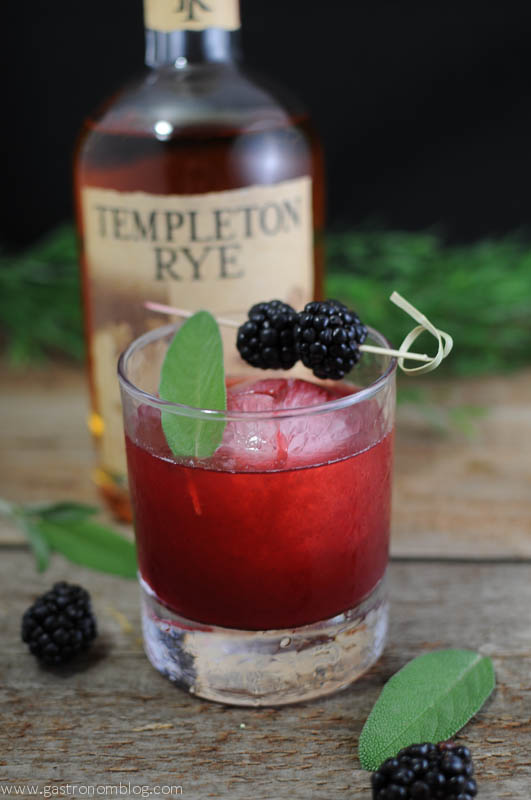 Sage Advice Cocktail with sage leaves and blackberries on cocktail pick. Rye Whiskey bottle on wooden background
