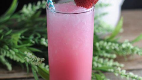 Strawberry Tropical Chiller - pink cocktail in tall glass with strawberry on rim and blue/white striped straw. Greenery and white pitcher in background