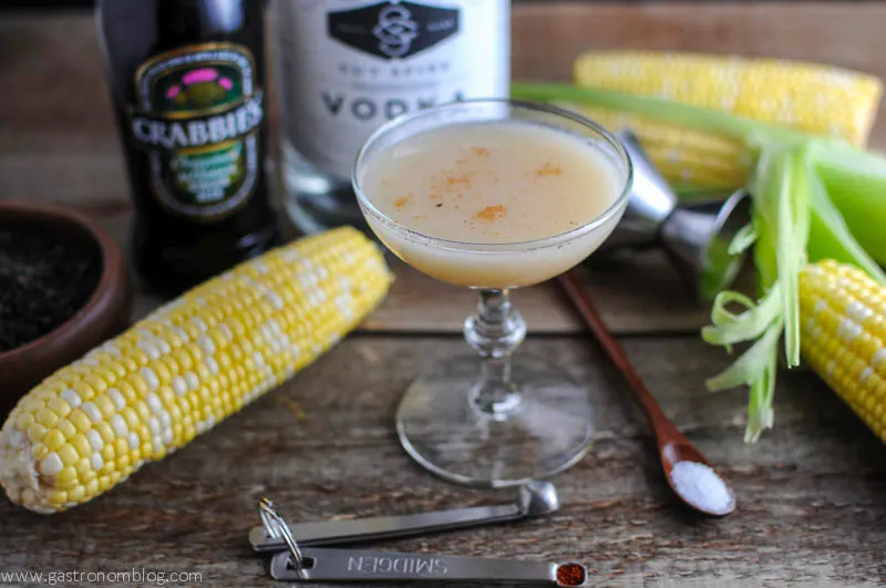 The Nebraskan - A Corn cocktail recipe with ginger beer, corn, sweet corn simple syrup, salt and pepper, touch of cayenne from Gastronomblog. This corn milk based drink is sweet and spicy. #cocktail #gastronomblog #saltandpepper #ginger #corn The Nebraskan Cocktail in a coupe with ears of corn, jigger and wooden spoon. Bottles of ginger beer and vodka in background