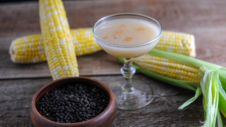 The Nebraskan - corn cocktail in coupe, wooden bowl of peppercorns and cobs of corn in background