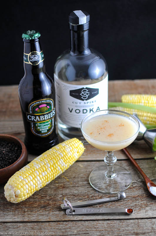 Yellow cocktail in coupe, cob of corn, ginger beer and vodka bottle behind, spoons and wooden spoon