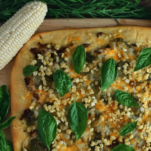 Pizza wtih corn and basil on cutting board top shot with cobs of corn and basil leaves