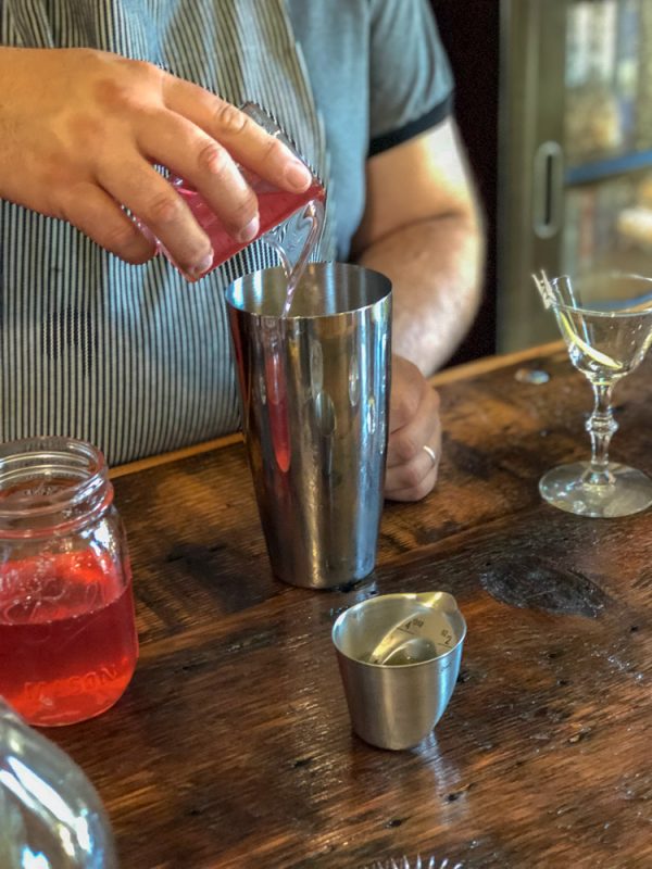 Rhubarb Sour cocktail syrup being poured into shaker. Jar, coupe and jigger on wood counter