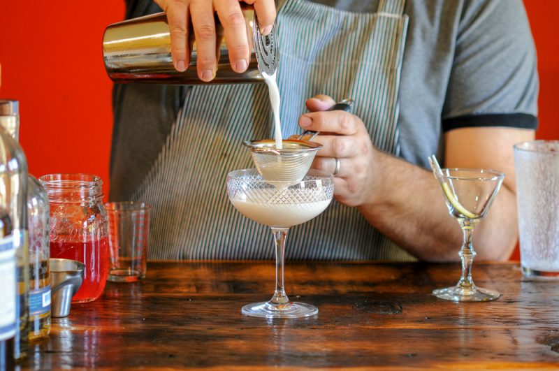 Rhubarb Sour cocktail being poured from shaker to a coupe. Jar of syrup, jigger and coupe on wooden counter