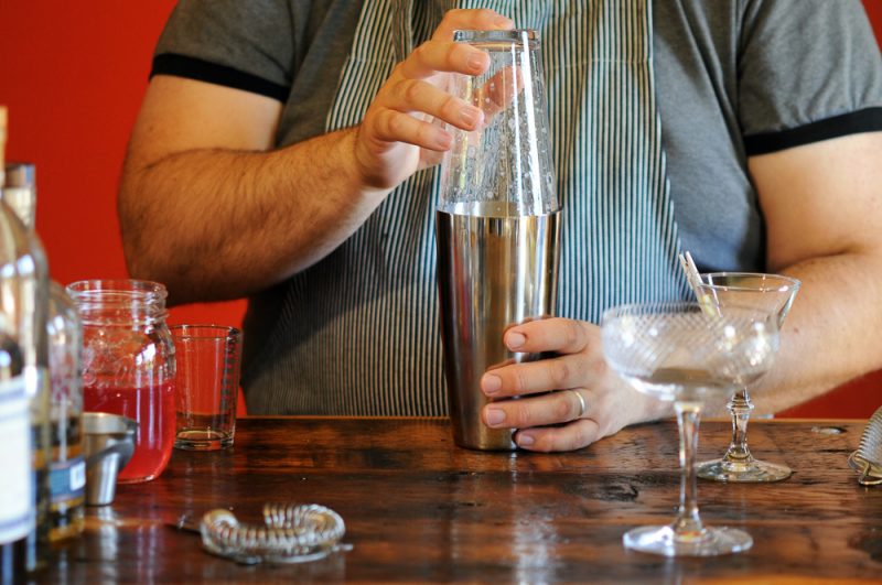 Rhubarb Sour cocktail being shaken in a glass and stainless shaker. Coupes, jigger and jar on counter
