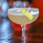 Yellow cocktail with egg white topper in a coupe, lemon peel pinned to side of glass