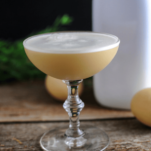 Brown cocktail in coupe with white foam, white bottle and eggs behind