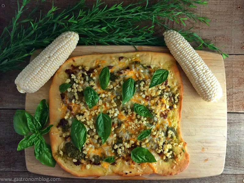 Pesto, Sausage and Corn Pizza - perfect for those fresh summer veggies from Gastronomblog. This corn pizza recipe is topped with fresh basil and corn from Nebraska. #recipe #cheese #corn #pizza #gastronomblog Sausage Corn and Pesto Pizza with ears of corn and basil on a cutting board