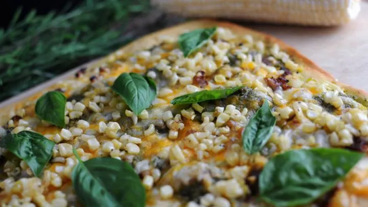 Pesto Corn Pizza on cutting board with corn cob in back. Pizza has basil leaves, corn, sausage and pesto.