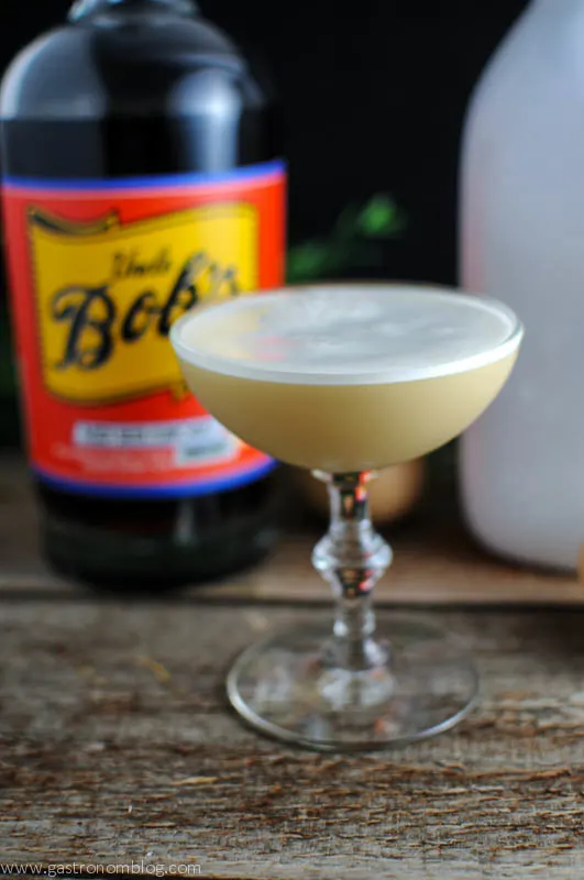 Tan Root Beer Whiskey cocktail in coupe with white foam, white bottle and eggs behind