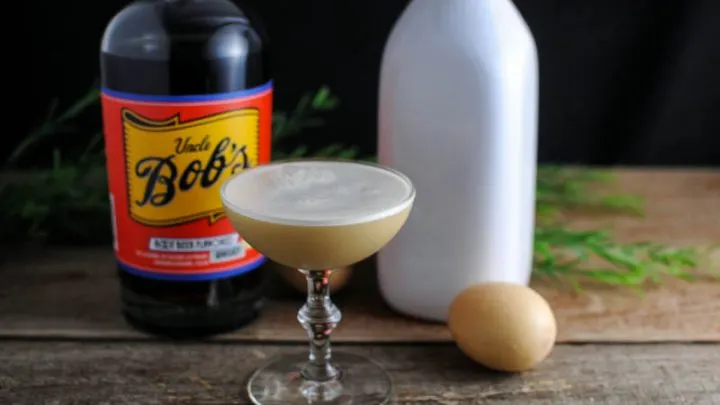 Tan Root Beer Whiskey cocktail in coupe with white foam, white bottle and eggs behind