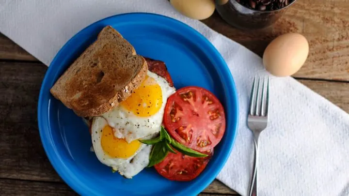 Summer Breakfast Sandwich - fried eggs on wheat toast with basil and tomato slices on blue plate. Plate is on white napkin with fork, eggs