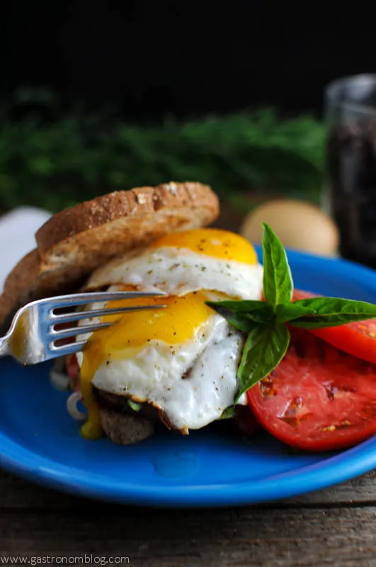 Fork cutting running egg on wheat bread with tomatoes on blue plate. Eggs and greenery in the background