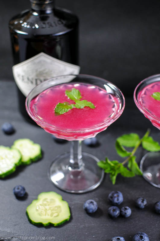 Pink cocktail in two coupes, mint leaves on top. Blueberries, cucumbers and mint, along with Hendrick's Gin bottle in background on slate